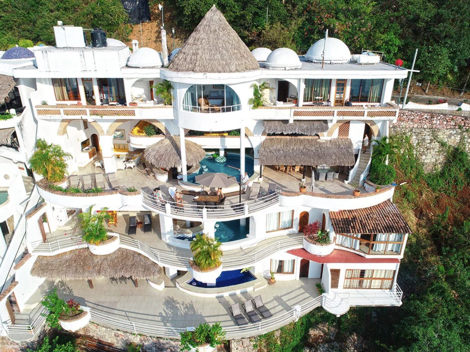 The villa from above: five levels of white terraces, palapa roofs, and pools stacked down a jungle hillside.